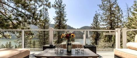 A deck with outdoor furniture, a table with a vase of flowers, and a view of a lake surrounded by trees.