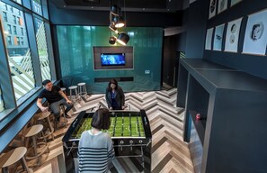 A modern indoor space with a foosball table, seating area, and a television screen displaying a program. - Student Only Verde Newcastle (Newcastle upon Tyne)