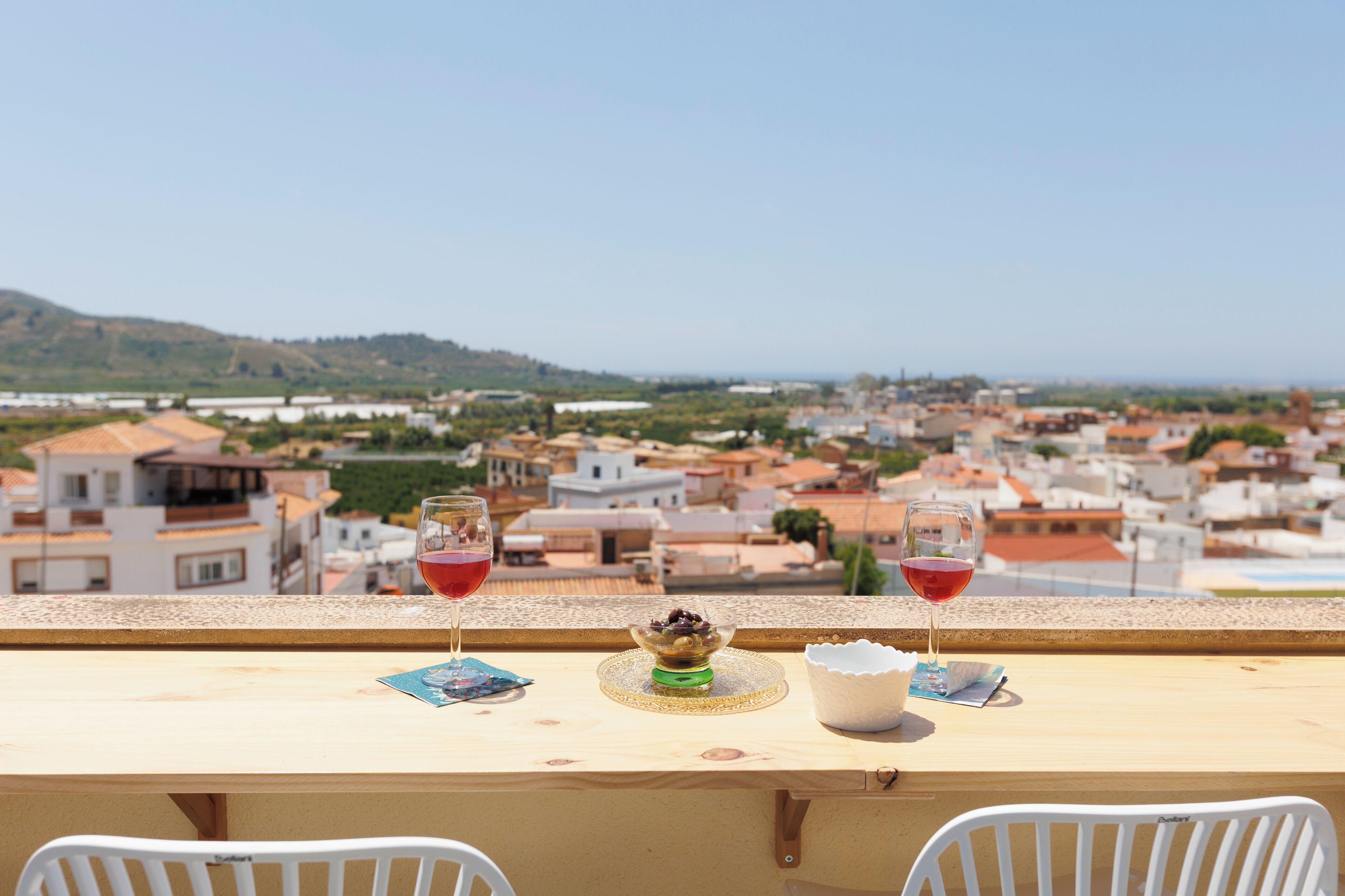 A balcony with a table, two glasses of red wine, a bowl of fruit, and a view of a town.