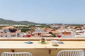 A balcony with a table, two glasses of red wine, a bowl of fruit, and a view of a town.