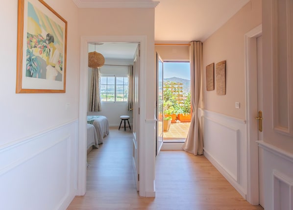 A hallway with a bed, a stool, and a view of an outdoor garden through an open door.
