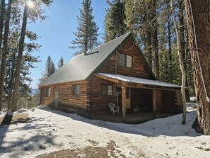 A log cabin with a porch and a chimney, surrounded by snow and trees. - Handcrafted Cozy Cabin Retreat with Scenic Mountain Views in McCall, Idaho (McCall)