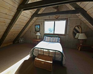 A bedroom with a sloped ceiling, a bed with a patterned bedspread, a bedside table with a lamp, a small round mirror, and a window with a view of trees. - Handcrafted Cozy Cabin Retreat with Scenic Mountain Views in McCall, Idaho (McCall)
