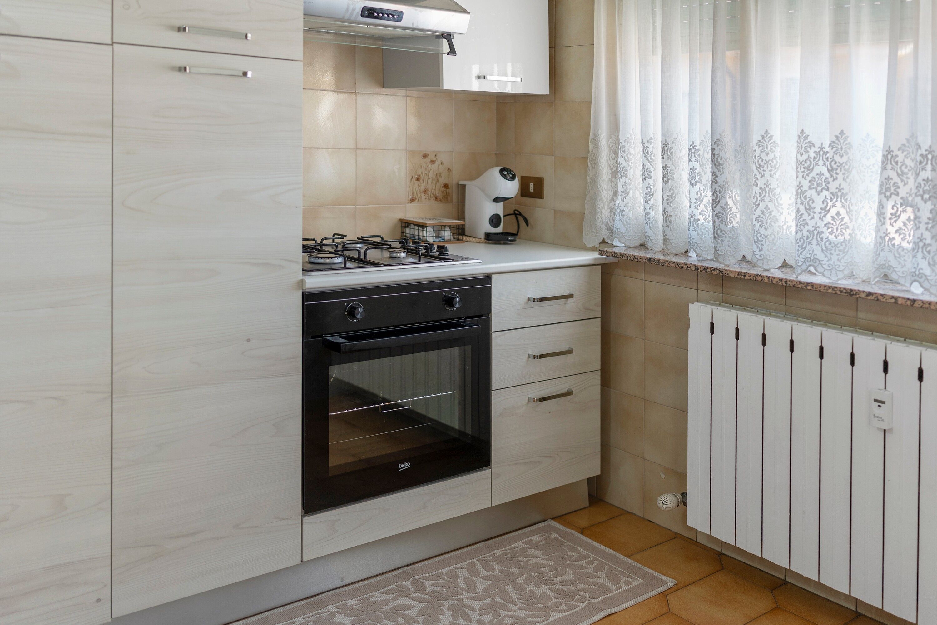 Eine Küche mit Gasherd, Backofen, weißen Schränken und einem Fenster mit durchsichtigen Vorhängen.