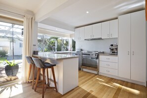 A modern kitchen with white cabinets, a central island, bar stools, and a view of greenery outside. - Moonah Sands with Cottage (Rye)