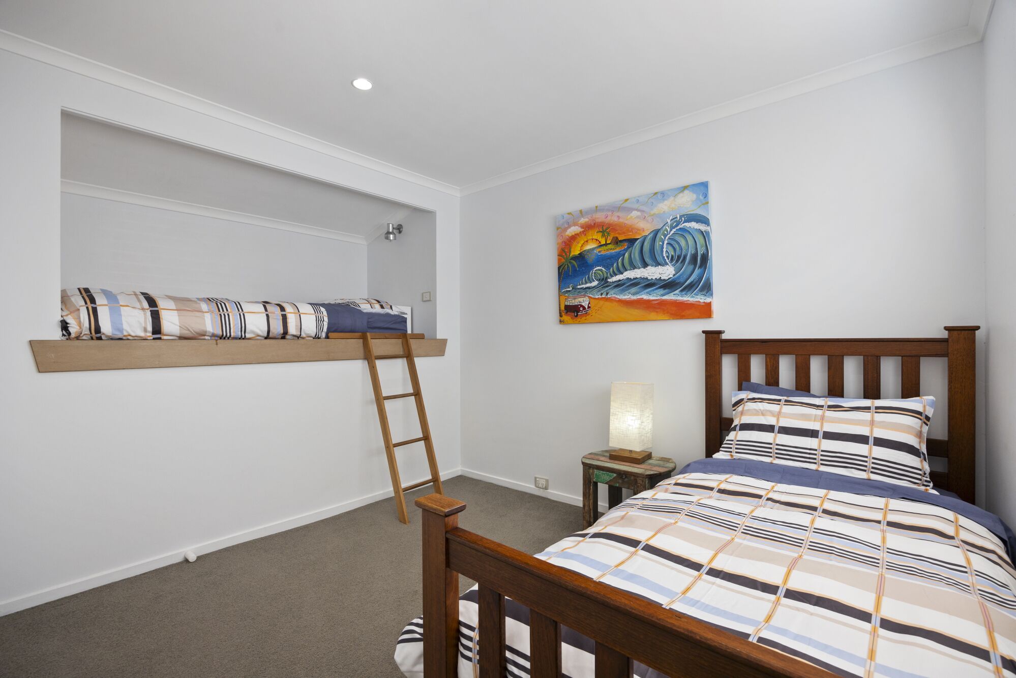 Un dormitorio con una cama litera, una escalera, una mesita de noche con lámpara y un cuadro en la pared.
