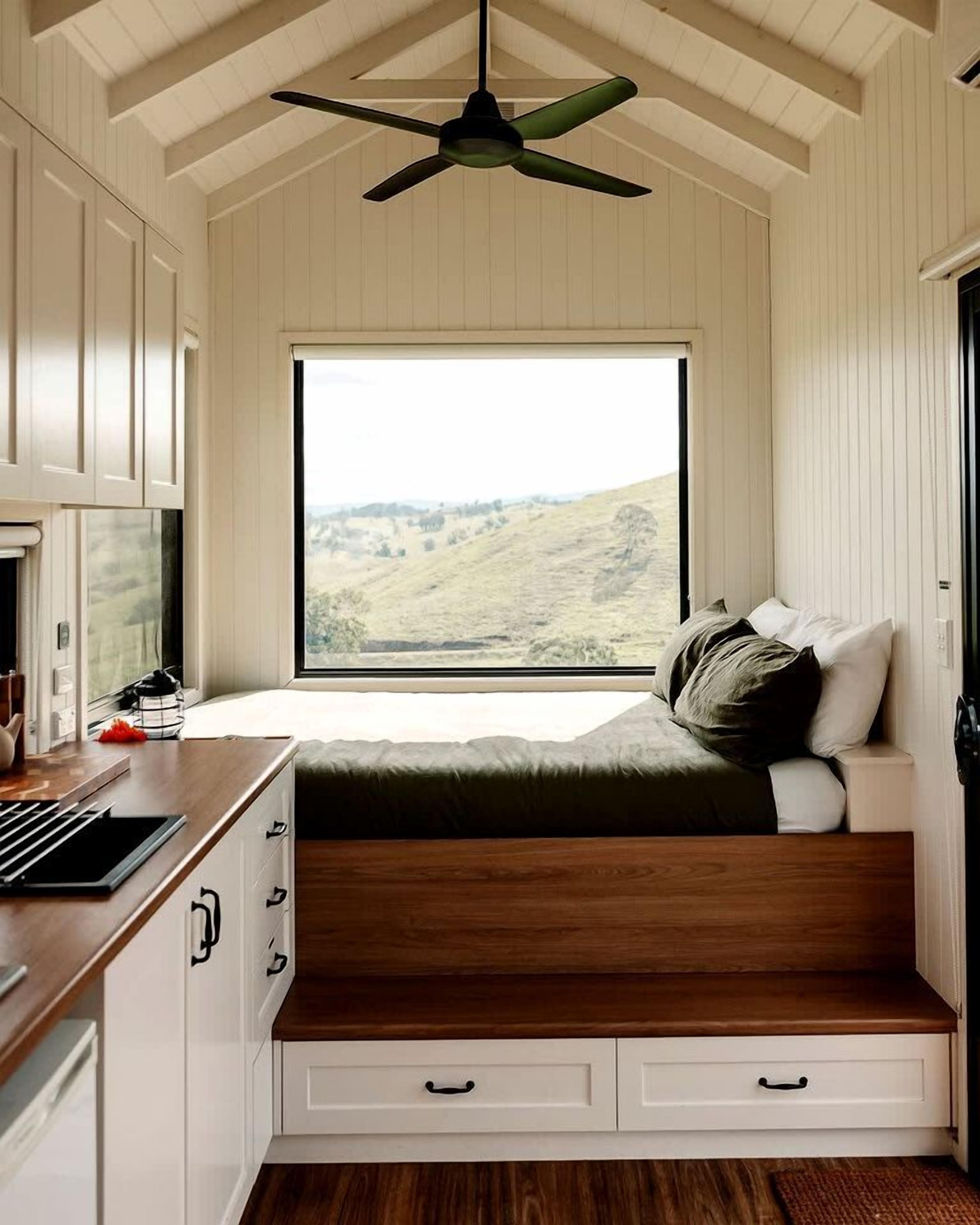A bedroom with a built-in bed, a window with a view of a hilly landscape, a ceiling fan, and a kitchenette area.