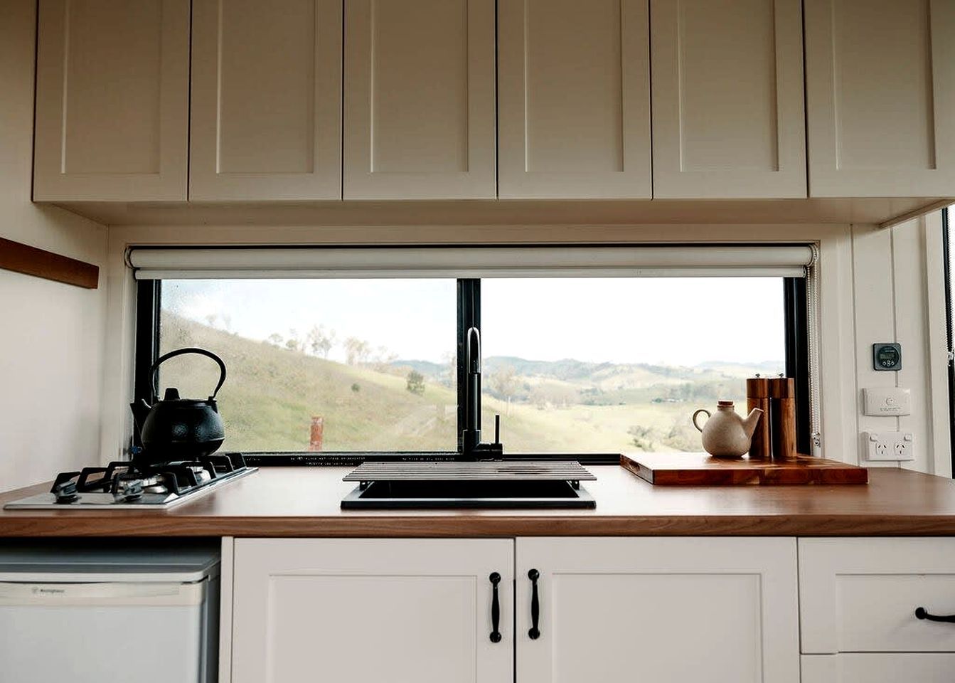 A kitchen with a gas stove, a kettle, and a view of a green landscape through a window.