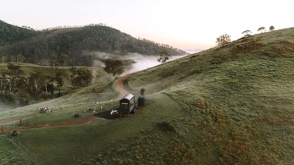 A rural landscape with a small building, a dirt path, and a body of water in the distance. - Mountain Tiny House with Spectacular Landscape Views in New South Wales (Bulliac)
