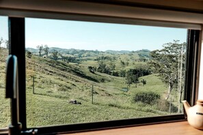 A view through a window of a rural landscape with rolling hills, trees, and a clear sky. - Mountain Tiny House with Spectacular Landscape Views in New South Wales (Bulliac)