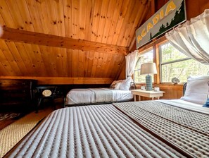 A cabin interior with a wooden ceiling, two beds with striped bedding, a nightstand with a lamp, and a window with curtains. - Charming Cabin with Fireplace Near Scenic Trails in Mount Desert, Maine (Mount Desert)