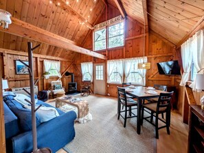 A wooden cabin interior with a sofa, dining table, chairs, a fireplace, and a TV. - Charming Cabin with Fireplace Near Scenic Trails in Mount Desert, Maine (Mount Desert)