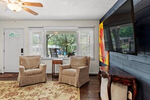 A living room with two beige armchairs, a wooden coffee table, a large TV mounted on a dark wall, and a window with blinds. - New- Minutes from Atl Attractions, Hiking, Beltline, Backyard, Grill,  Pet Friendly (Atlanta)