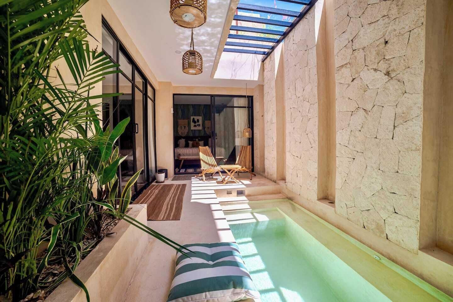 A sunlit indoor pool area with a lounge chair, a small table, and a potted plant.