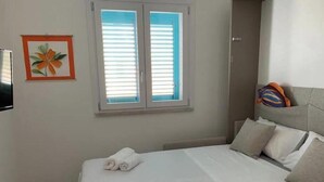 Habitación de hotel con cama, dos toallas enrolladas, ventana con persianas, un cuadro enmarcado y almohada con un diseño colorido.
