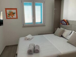 Ein Hotelzimmer mit einem Bett, zwei zusammengerollten Handtüchern, einem Fenster mit Jalousien, einem gerahmten Bild und einem Kissen mit farbigem Muster.