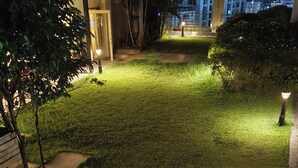 A well-lit garden pathway with stepping stones leading to a door, surrounded by lush greenery and a cityscape in the background. - Cityscape, Siddha Xanadu, Newtown: Gym, pool, restaurant, garden, 24/7 security. (Kolkata)