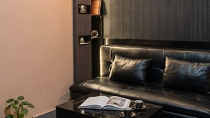 A modern living room with a black leather sofa, a black coffee table with a book and a cup, a potted plant, and a wall-mounted shelf with decorative items. - Cityscape, Siddha Xanadu, Newtown: Gym, pool, restaurant, garden, 24/7 security. (Kolkata)