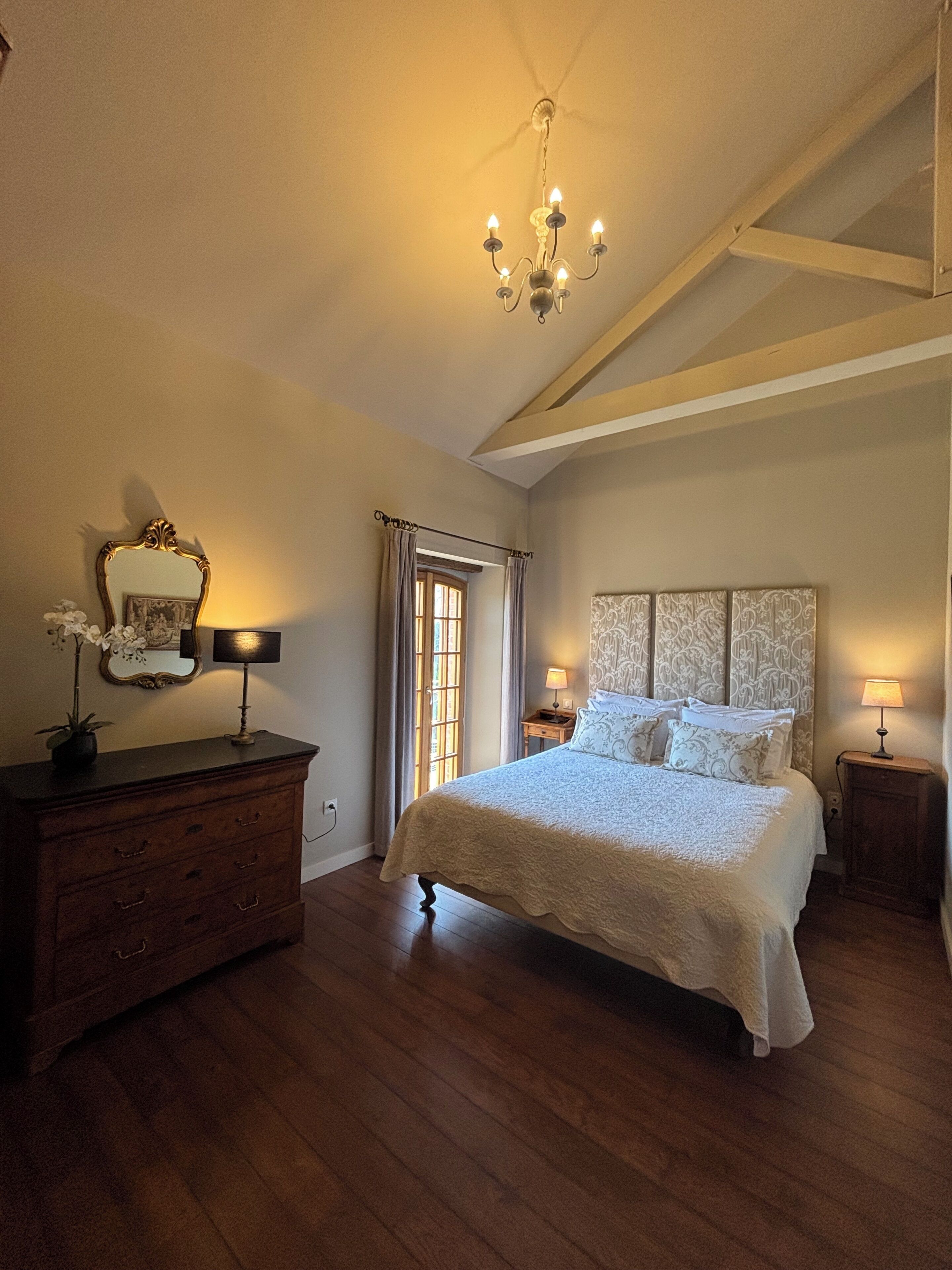 Een slaapkamer met een groot bed, een dressoir, een spiegel, een kroonluchter en een raam met gordijnen.