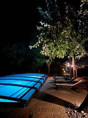 Zona de piscina exterior amb una piscina il·luminada de blau, gandules i una pèrgola amb ombrelles.