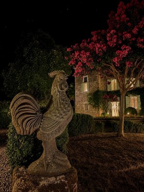 Una estàtua de gall de pedra en un jardí, amb un edifici i arbres florits al fons.