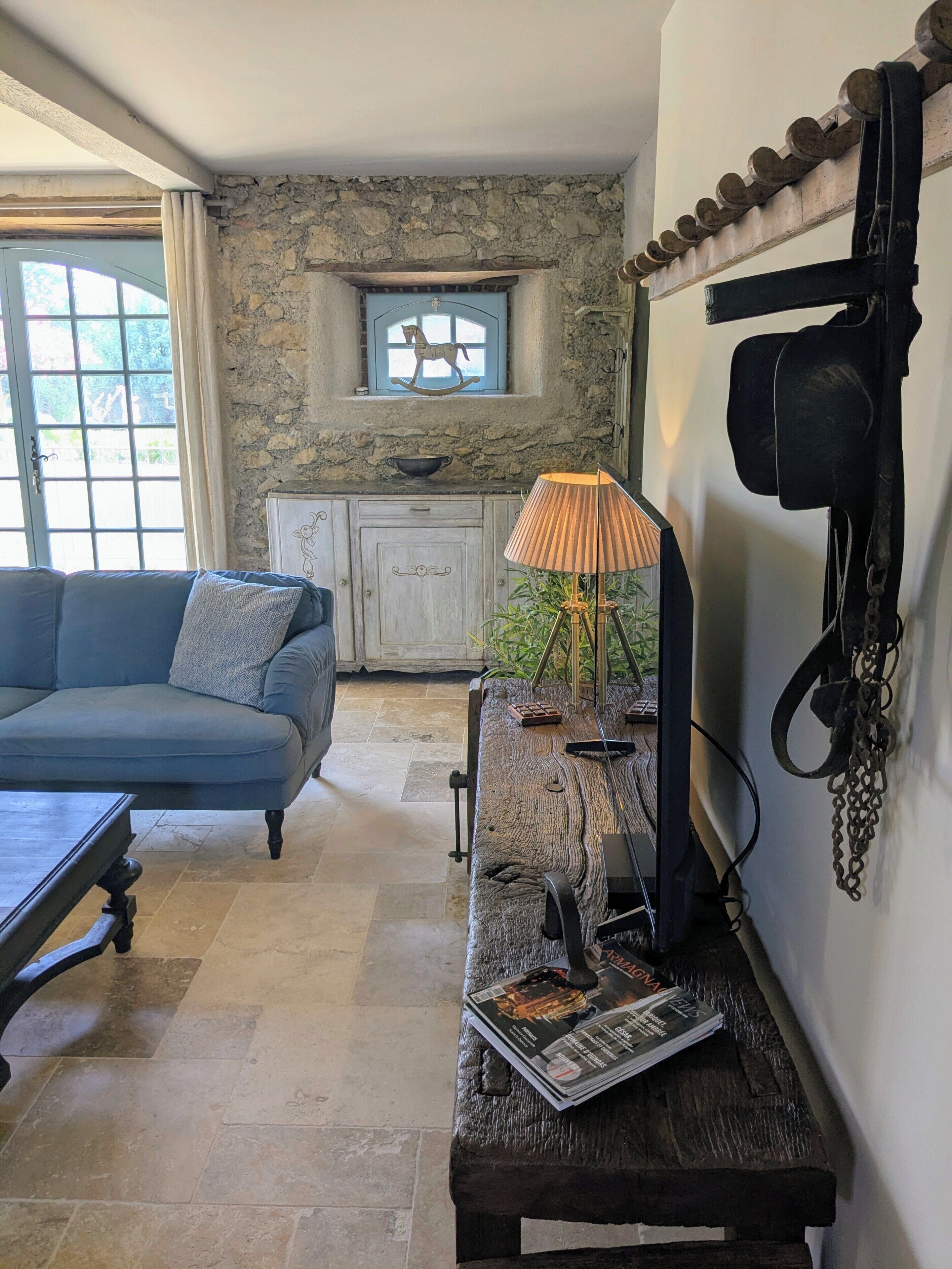 Une pièce avec un canapé bleu, un banc en bois avec des magazines, une lampe, un mur en pierre, une fenêtre avec un cheval à bascule et un meuble blanc.