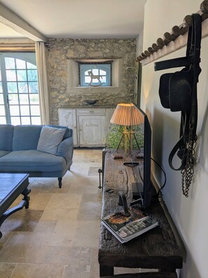 A room with a blue sofa, a wooden bench with magazines, a lamp, a stone wall, a window with a rocking horse, and a white cabinet. - Luxury chateau retreat; pool, summer kitchen, outdoor cinema & Madiran vineyards (Castelnau-Rivière-Basse)