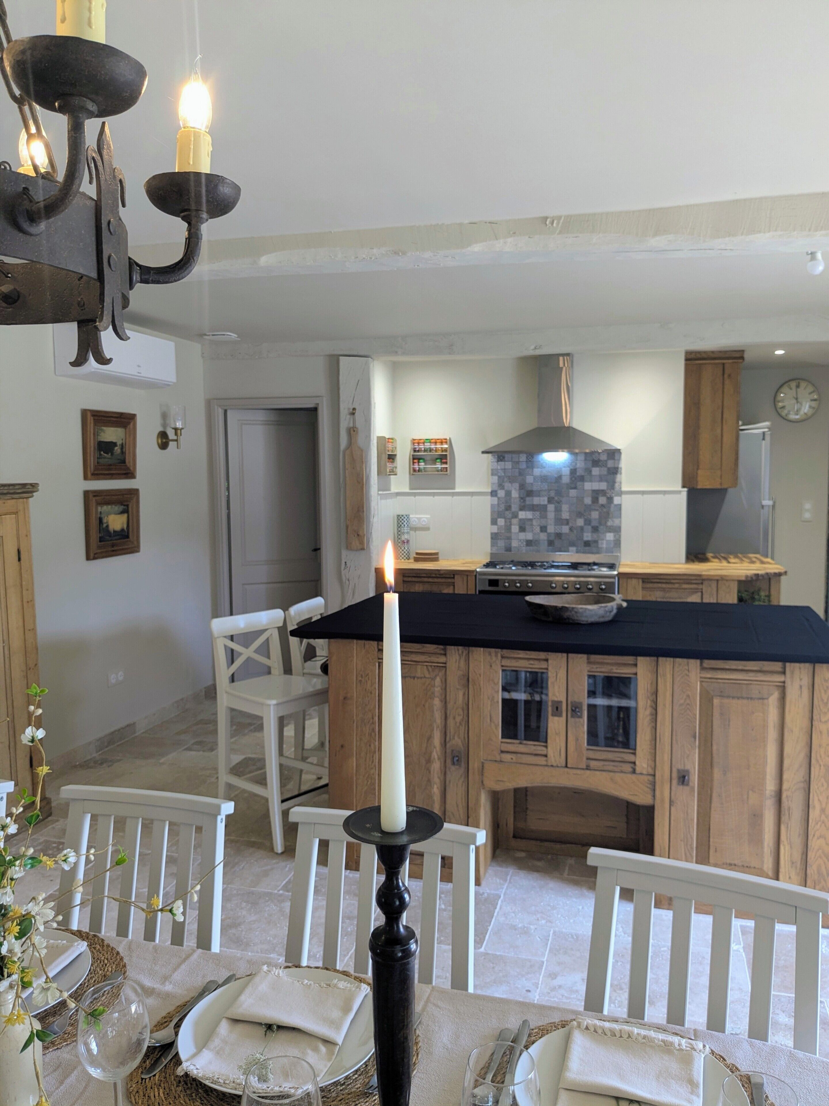 A dining area with a table set for two, a candle centerpiece, and a kitchen in the background with wooden cabinets and a tiled backsplash.