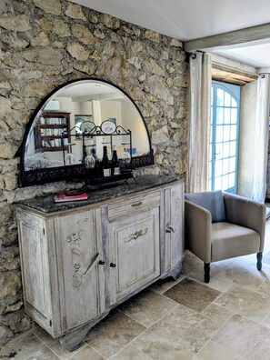 A stone wall with a large mirror above a wooden cabinet. There is a chair to the right and a bookshelf in the background. - Luxury chateau retreat; pool, summer kitchen, outdoor cinema & Madiran vineyards (Castelnau-Rivière-Basse)