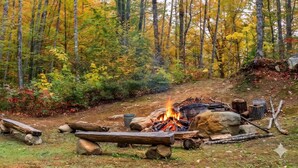 A forest campsite with a fire, logs, rocks, and a metal bucket. - White Mountains A-Frame | Off-Grid, Stars, Firepit (Stoneham)