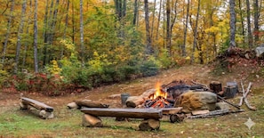 A forest campsite with a fire, logs, rocks, and a metal bucket. - White Mountains A-Frame | Off-Grid, Stars, Firepit (Stoneham)