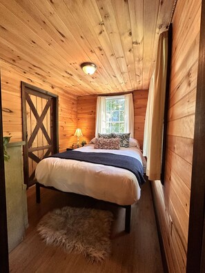A wooden cabin bedroom with a bed, a window with curtains, a lamp, and a door.