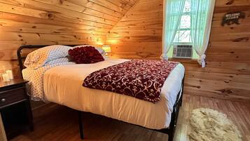 Un dormitorio tipo cabaña de madera con una cama, una mesita de noche, una alfombra de piel y una ventana con cortinas.