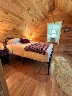 Un dormitorio tipo cabaña de madera con una cama, una mesita de noche, una alfombra de piel y una ventana con cortinas.