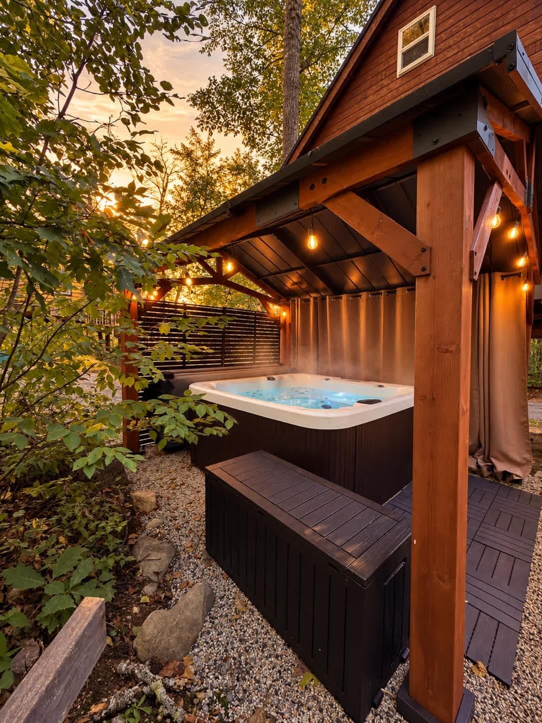 A hot tub under a wooden pergola with string lights, surrounded by a garden with rocks and foliage.