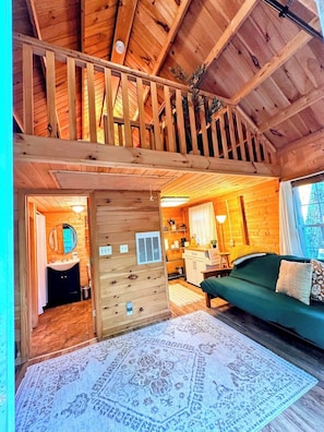 A wooden interior with a loft bedroom, a green sofa, and a patterned rug. - Boutique Treehouse HOT TUB Firepit Trails & Views! (Stoneham)