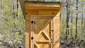 Uma casa de banho de madeira com porta em forma de cruz, situada em terreno rochoso com árvores ao fundo.