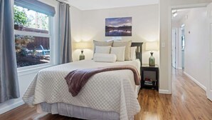 Quarto com uma cama grande, duas mesas de cabeceira, uma janela com cortinas, um quadro na parede e um corredor.