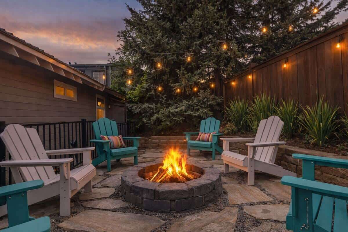 A fire pit surrounded by Adirondack chairs, with a wooden fence and string lights in the background.