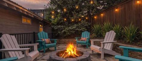 A fire pit surrounded by Adirondack chairs, with a wooden fence and string lights in the background.