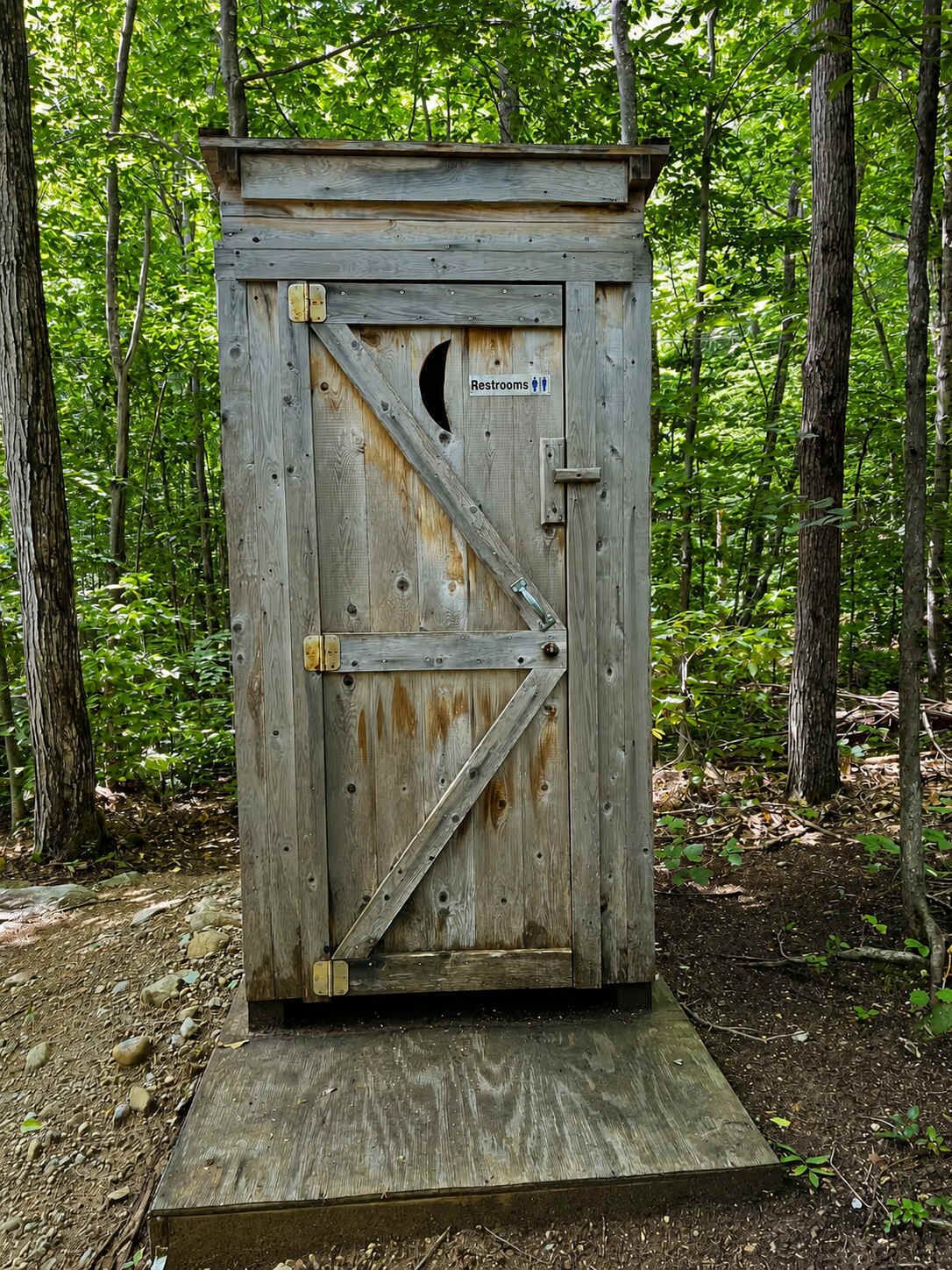 A rustic wooden outhouse with a crescent moon cutout on the door, labeled 'Restrooms,' set in a forested area.