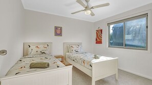 A bedroom with two white beds, a ceiling fan, a window with a view, and a wall decoration.