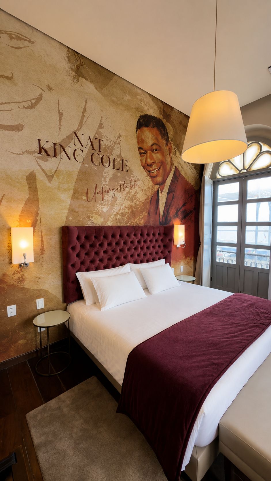 Quarto de hotel com uma cama grande, cabeceira vermelha, um mural na parede e uma janela com vista.