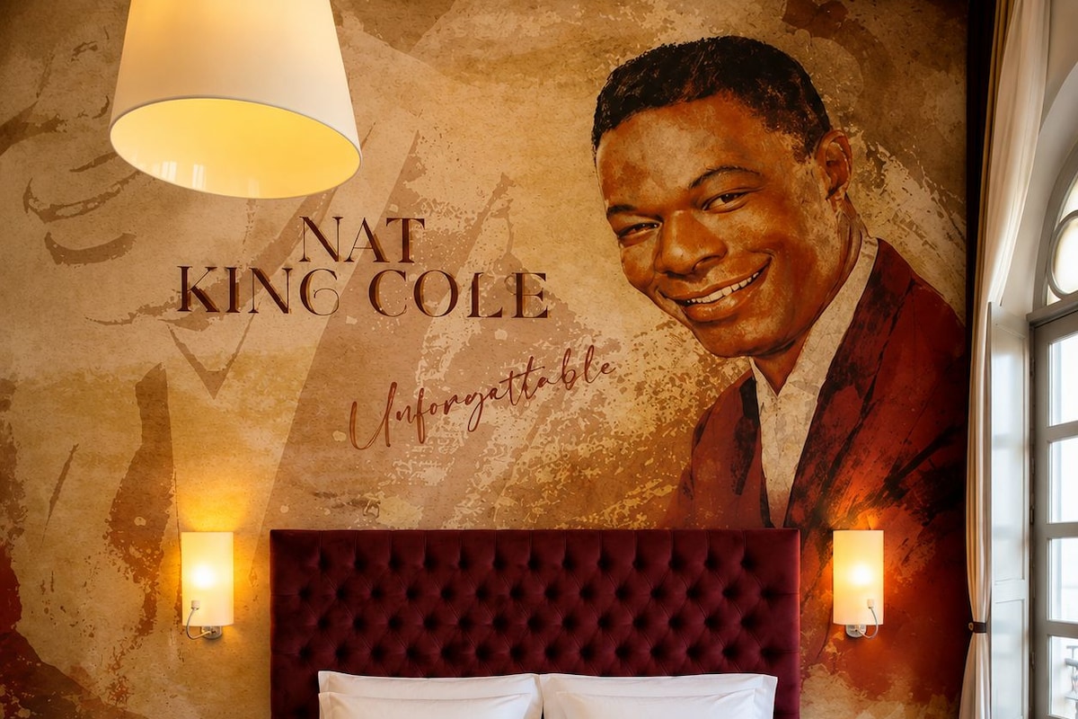 Quarto de hotel com uma cama grande, cabeceira estofada, duas luminárias de mesa e um mural na parede.