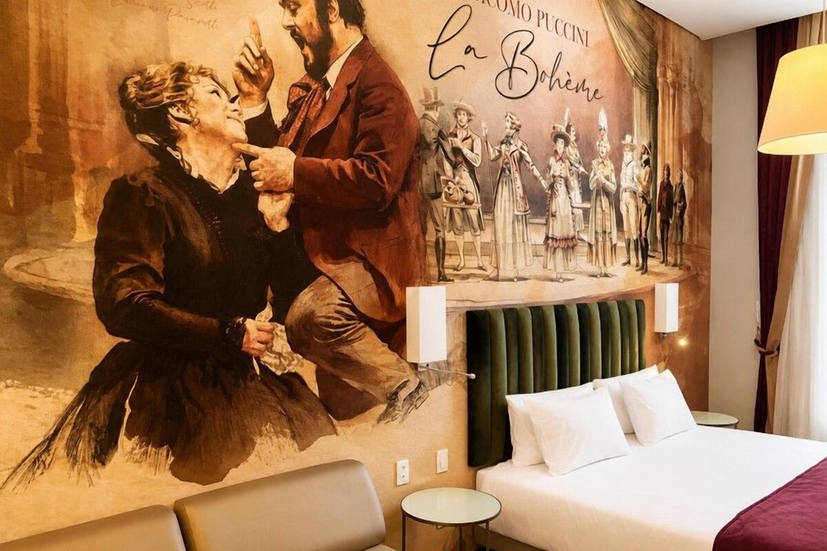 Quarto de hotel com um grande mural, uma cama com lençóis brancos, cabeceira verde e uma pequena mesa de cabeceira.