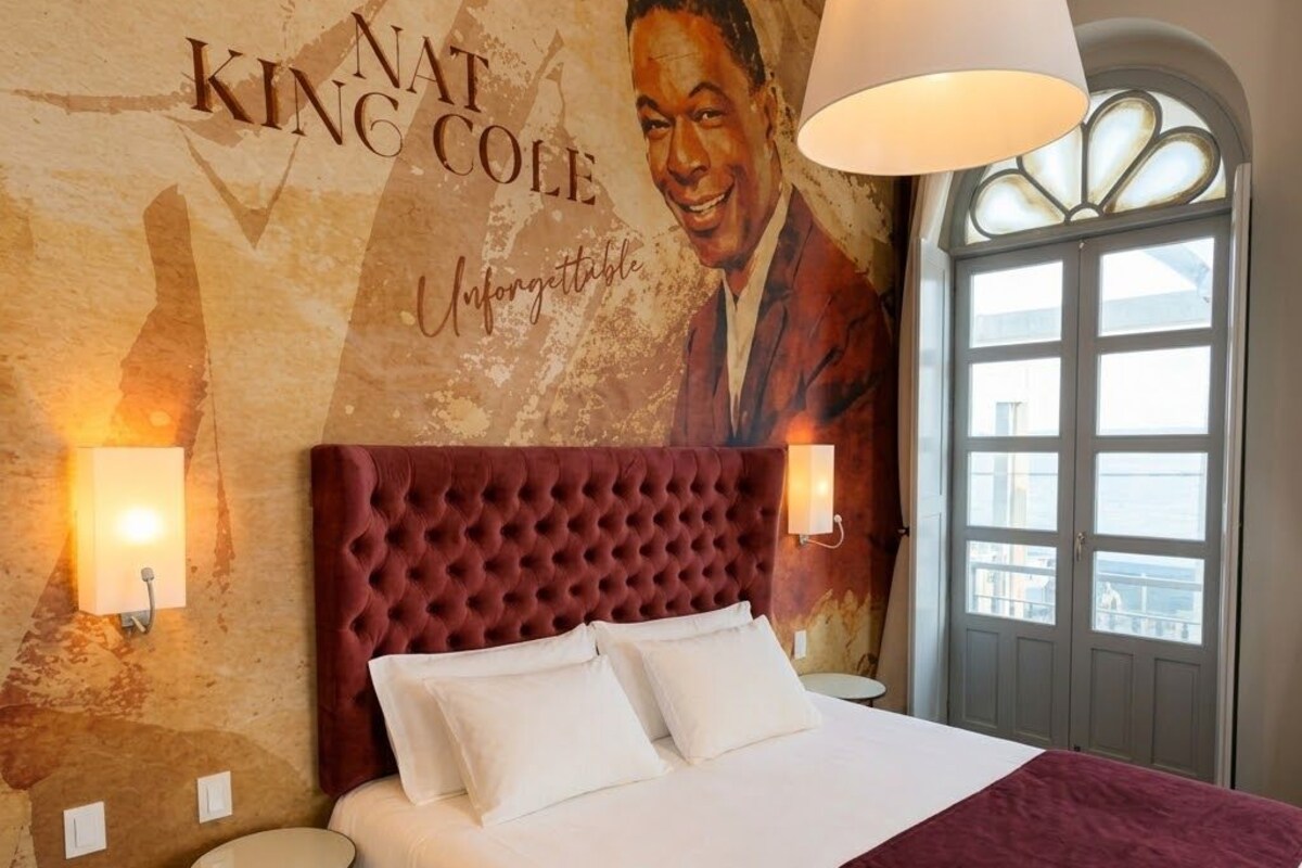 Quarto de hotel com uma cama grande, cabeceira estofada, duas luminárias de apoio e um mural na parede.