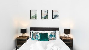 Een net opgemaakt bed met witte lakens en twee kussens, met aan weerszijden nachtkastjes met lampen, en daarboven drie kunstwerken in lijsten aan de muur.