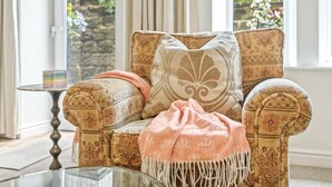 Un fauteuil à motifs, agrémenté d’un coussin et d’un plaid, placé près d’une table en verre et d’une petite table d’appoint sur laquelle se trouve une bougie.
