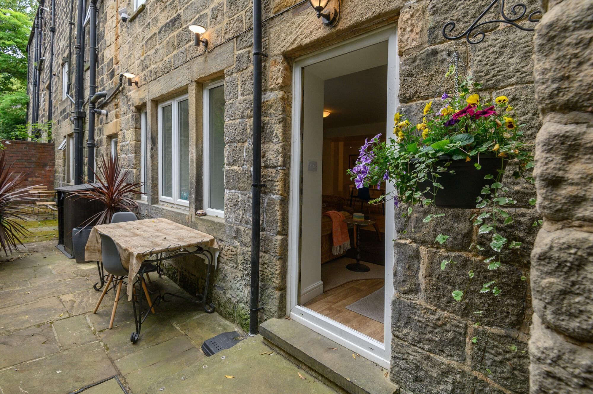 A stone building with a wooden table and chairs outside, a hanging flower basket, and a door leading inside.