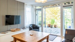 Ett modernt vardagsrum med en platt-TV, ett soffbord i trä, en lädersoffa och en skjutdörr i glas som leder ut till en trädgård.